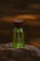 A green perfume bottle with a wooden cap placed on a textured rock surface. The background is blurred with warm amber lighting, creating a natural and earthy ambiance.