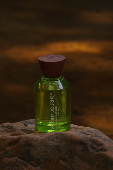 A green perfume bottle with a wooden cap placed on a textured rock surface. The background is blurred with warm amber lighting, creating a natural and earthy ambiance.