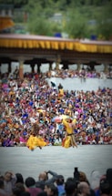 Two dancers in traditional attire perform energetically in front of a large, colorful crowd. The background features a structure adorned with yellow and orange decorations. The audience is dressed in vibrant clothing, creating a lively and festive atmosphere.