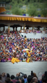 Two dancers in traditional attire perform energetically in front of a large, colorful crowd. The background features a structure adorned with yellow and orange decorations. The audience is dressed in vibrant clothing, creating a lively and festive atmosphere.