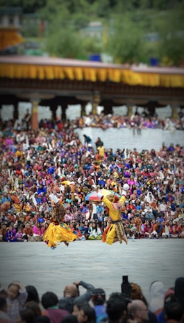 Two dancers in traditional attire perform energetically in front of a large, colorful crowd. The background features a structure adorned with yellow and orange decorations. The audience is dressed in vibrant clothing, creating a lively and festive atmosphere.