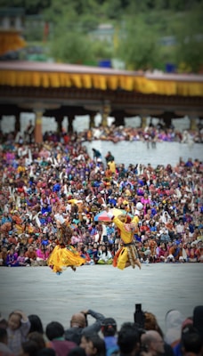 Two dancers in traditional attire perform energetically in front of a large, colorful crowd. The background features a structure adorned with yellow and orange decorations. The audience is dressed in vibrant clothing, creating a lively and festive atmosphere.