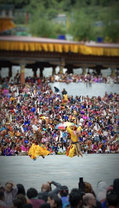 Two dancers in traditional attire perform energetically in front of a large, colorful crowd. The background features a structure adorned with yellow and orange decorations. The audience is dressed in vibrant clothing, creating a lively and festive atmosphere.