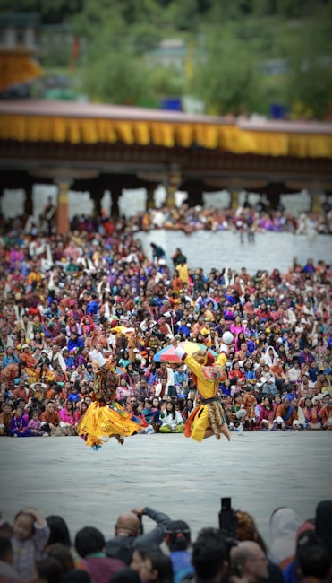 Two dancers in traditional attire perform energetically in front of a large, colorful crowd. The background features a structure adorned with yellow and orange decorations. The audience is dressed in vibrant clothing, creating a lively and festive atmosphere.