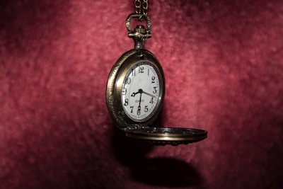 A vintage pocket watch is suspended against a rich, red velvet background. The watch is open, revealing a white clock face with black numerals and hands. The metallic case of the watch has an antique look, adding to its timeless charm.