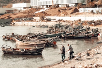 Several small wooden fishing boats are docked along a calm shoreline. Two people are interacting near the water's edge, possibly discussing or handling something. The background features industrial buildings, a sloped, earthy terrain, and sparse vegetation.