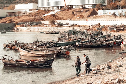 Several small wooden fishing boats are docked along a calm shoreline. Two people are interacting near the water's edge, possibly discussing or handling something. The background features industrial buildings, a sloped, earthy terrain, and sparse vegetation.