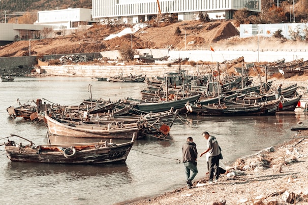 Several small wooden fishing boats are docked along a calm shoreline. Two people are interacting near the water's edge, possibly discussing or handling something. The background features industrial buildings, a sloped, earthy terrain, and sparse vegetation.
