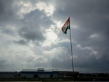 A tall flagpole holds an Indian flag, prominently displayed under a cloudy sky with visible patches of sunlight breaking through. The scene is set against an industrial backdrop with a large building on the horizon.