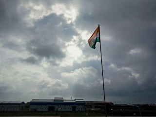 A tall flagpole holds an Indian flag, prominently displayed under a cloudy sky with visible patches of sunlight breaking through. The scene is set against an industrial backdrop with a large building on the horizon.
