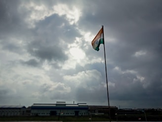 A tall flagpole holds an Indian flag, prominently displayed under a cloudy sky with visible patches of sunlight breaking through. The scene is set against an industrial backdrop with a large building on the horizon.