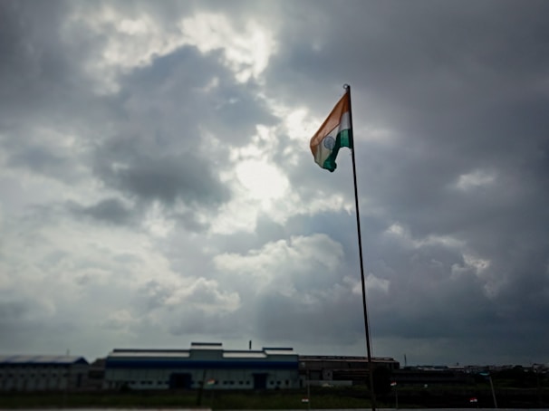 A tall flagpole holds an Indian flag, prominently displayed under a cloudy sky with visible patches of sunlight breaking through. The scene is set against an industrial backdrop with a large building on the horizon.