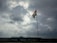 A tall flagpole holds an Indian flag, prominently displayed under a cloudy sky with visible patches of sunlight breaking through. The scene is set against an industrial backdrop with a large building on the horizon.