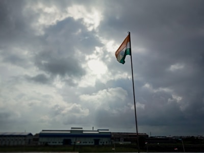 A tall flagpole holds an Indian flag, prominently displayed under a cloudy sky with visible patches of sunlight breaking through. The scene is set against an industrial backdrop with a large building on the horizon.