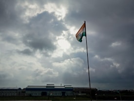 A tall flagpole holds an Indian flag, prominently displayed under a cloudy sky with visible patches of sunlight breaking through. The scene is set against an industrial backdrop with a large building on the horizon.