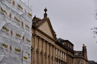 A historic building with classical architectural features is partially covered by scaffolding and protective sheeting with the logo 'Young's Roofing Limited'. The building facade features columns and intricate stonework, characteristic of traditional styles. The sky is overcast, giving the scene a somewhat subdued appearance.