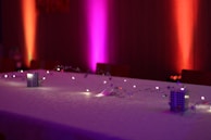 A long table is decorated with delicate string lights and small decorative objects. The background features colorful lighting with a gradient of pink, purple, and orange hues on the walls, creating a cozy and inviting atmosphere.