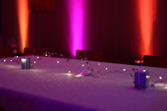 A long table is decorated with delicate string lights and small decorative objects. The background features colorful lighting with a gradient of pink, purple, and orange hues on the walls, creating a cozy and inviting atmosphere.