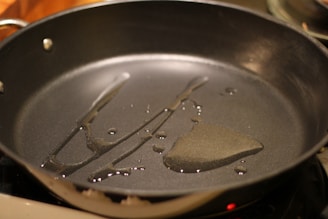 A black frying pan sits on a stovetop with a small amount of cooking oil spread across its surface. The oil glistens under the light, creating a shiny and reflective texture. The pan's handle and the edge of the stovetop can be seen, suggesting it's ready for cooking.