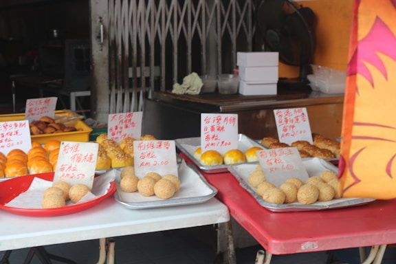 A marketplace or stall displaying various types of pastries and bread on several trays. The baked goods are arranged neatly, each with a label written in Chinese and prices in RM. The backdrop includes a metal gate and some kitchen equipment.