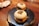 Two steamed buns with a filling, presented on a black, textured plate. The buns have some sort of marking or character on top of each, potentially suggesting a specific flavor or restaurant brand. They are neatly arranged on a wooden table, with some paper napkins and part of a spoon visible in the background, creating a cozy dining setting.
