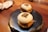 Two steamed buns with a filling, presented on a black, textured plate. The buns have some sort of marking or character on top of each, potentially suggesting a specific flavor or restaurant brand. They are neatly arranged on a wooden table, with some paper napkins and part of a spoon visible in the background, creating a cozy dining setting.