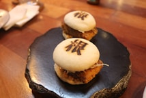 Two steamed buns with a filling, presented on a black, textured plate. The buns have some sort of marking or character on top of each, potentially suggesting a specific flavor or restaurant brand. They are neatly arranged on a wooden table, with some paper napkins and part of a spoon visible in the background, creating a cozy dining setting.