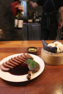 A wooden table setting with a plate of sliced roasted duck garnished with herbs and a rich sauce. Next to it, a bamboo steamer basket contains steamed buns. In the background, there are two blurred figures, one holding wine bottles.