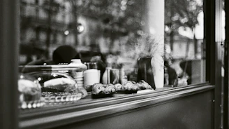 A black and white photograph captures the interior of a shop window displaying various baked goods, including croissants. The scene includes a glass cake stand, a stack of cups, and some decorative elements like a vase with wheat-like plants. Reflections in the glass reveal a blurred, leafy background with indistinct buildings.