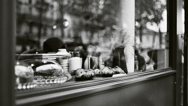 A black and white photograph captures the interior of a shop window displaying various baked goods, including croissants. The scene includes a glass cake stand, a stack of cups, and some decorative elements like a vase with wheat-like plants. Reflections in the glass reveal a blurred, leafy background with indistinct buildings.