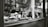 A black and white photograph captures the interior of a shop window displaying various baked goods, including croissants. The scene includes a glass cake stand, a stack of cups, and some decorative elements like a vase with wheat-like plants. Reflections in the glass reveal a blurred, leafy background with indistinct buildings.