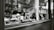 A black and white photograph captures the interior of a shop window displaying various baked goods, including croissants. The scene includes a glass cake stand, a stack of cups, and some decorative elements like a vase with wheat-like plants. Reflections in the glass reveal a blurred, leafy background with indistinct buildings.