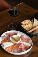 A wooden table displaying a plate of assorted charcuterie including prosciutto, salami, and cheese alongside a small dish of honey with a cinnamon stick. Next to the plate is a basket with slices of rustic bread. A glass of red wine is placed beside the basket, completing the setting.