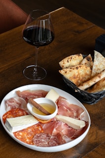 A wooden table displaying a plate of assorted charcuterie including prosciutto, salami, and cheese alongside a small dish of honey with a cinnamon stick. Next to the plate is a basket with slices of rustic bread. A glass of red wine is placed beside the basket, completing the setting.