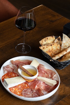 A wooden table displaying a plate of assorted charcuterie including prosciutto, salami, and cheese alongside a small dish of honey with a cinnamon stick. Next to the plate is a basket with slices of rustic bread. A glass of red wine is placed beside the basket, completing the setting.