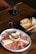 A wooden table displaying a plate of assorted charcuterie including prosciutto, salami, and cheese alongside a small dish of honey with a cinnamon stick. Next to the plate is a basket with slices of rustic bread. A glass of red wine is placed beside the basket, completing the setting.