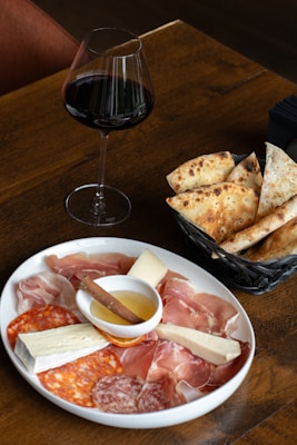 A wooden table displaying a plate of assorted charcuterie including prosciutto, salami, and cheese alongside a small dish of honey with a cinnamon stick. Next to the plate is a basket with slices of rustic bread. A glass of red wine is placed beside the basket, completing the setting.