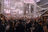 A bustling scene of a crowded holiday market with decorated stalls lined up, adorned with festive lights and garlands. The area is filled with people milling around, some holding drinks and food. Above, twinkling lights create a canopy-like effect, adding to the festive atmosphere. A modern building with large windows and the words 'Tower Plaza' can be seen in the background.
