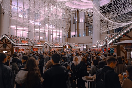 A bustling scene of a crowded holiday market with decorated stalls lined up, adorned with festive lights and garlands. The area is filled with people milling around, some holding drinks and food. Above, twinkling lights create a canopy-like effect, adding to the festive atmosphere. A modern building with large windows and the words 'Tower Plaza' can be seen in the background.