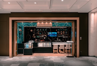 A small, modern café or bar with an elegant interior featuring teal and black decor. Glass displays showcase products, and shelves are lined with bottles and small items. Two people wearing face masks are behind the counter. Ambient lighting sets a cozy atmosphere, and there are a few chairs in front of the counter.