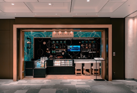 A small, modern café or bar with an elegant interior featuring teal and black decor. Glass displays showcase products, and shelves are lined with bottles and small items. Two people wearing face masks are behind the counter. Ambient lighting sets a cozy atmosphere, and there are a few chairs in front of the counter.