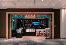A small, modern caf&eacute; or bar with an elegant interior featuring teal and black decor. Glass displays showcase products, and shelves are lined with bottles and small items. Two people wearing face masks are behind the counter. Ambient lighting sets a cozy atmosphere, and there are a few chairs in front of the counter.