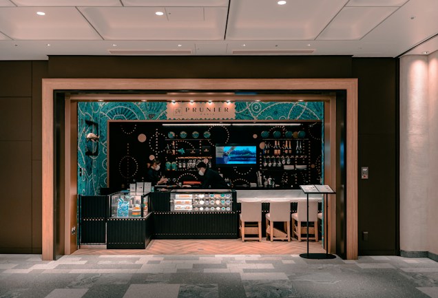 A small, modern caf&eacute; or bar with an elegant interior featuring teal and black decor. Glass displays showcase products, and shelves are lined with bottles and small items. Two people wearing face masks are behind the counter. Ambient lighting sets a cozy atmosphere, and there are a few chairs in front of the counter.