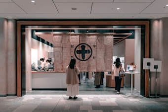 A modern shop interior with large fabric curtains hanging at the entrance featuring traditional symbols. Several people are visible inside, attending to various activities, and there are counters with objects and displays. The overall ambiance suggests a blend of traditional and contemporary styles.