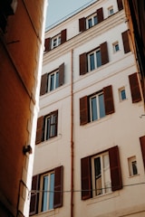 A tall building with light-colored walls and numerous windows featuring dark wooden shutters. The image captures an upward view from a narrow alley, highlighting the building's architectural details and clean lines. The sun casts shadows that add depth and contrast to the scene.