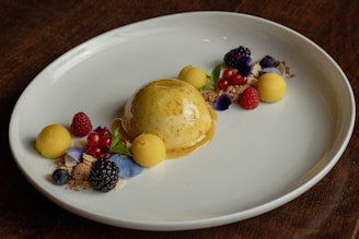 A visually appealing dessert on a white plate featuring a smooth, light brown dome of caramel or butterscotch panna cotta. Garnished with fresh fruit such as raspberries, blackberries, blueberries, and currants, alongside delicate yellow spheres that look like mango or passionfruit sorbet. Edible flowers and mint leaves add a touch of color and elegance, with crumbled cookie or granola bits scattered around for texture.