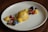 A visually appealing dessert on a white plate featuring a smooth, light brown dome of caramel or butterscotch panna cotta. Garnished with fresh fruit such as raspberries, blackberries, blueberries, and currants, alongside delicate yellow spheres that look like mango or passionfruit sorbet. Edible flowers and mint leaves add a touch of color and elegance, with crumbled cookie or granola bits scattered around for texture.