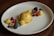 A visually appealing dessert on a white plate featuring a smooth, light brown dome of caramel or butterscotch panna cotta. Garnished with fresh fruit such as raspberries, blackberries, blueberries, and currants, alongside delicate yellow spheres that look like mango or passionfruit sorbet. Edible flowers and mint leaves add a touch of color and elegance, with crumbled cookie or granola bits scattered around for texture.