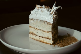 A slice of layered cake with a fluffy white frosting on top, sprinkled with cocoa powder, placed on a white plate. The cake has multiple layers with visible light brown filling, giving it a rich, textured appearance.