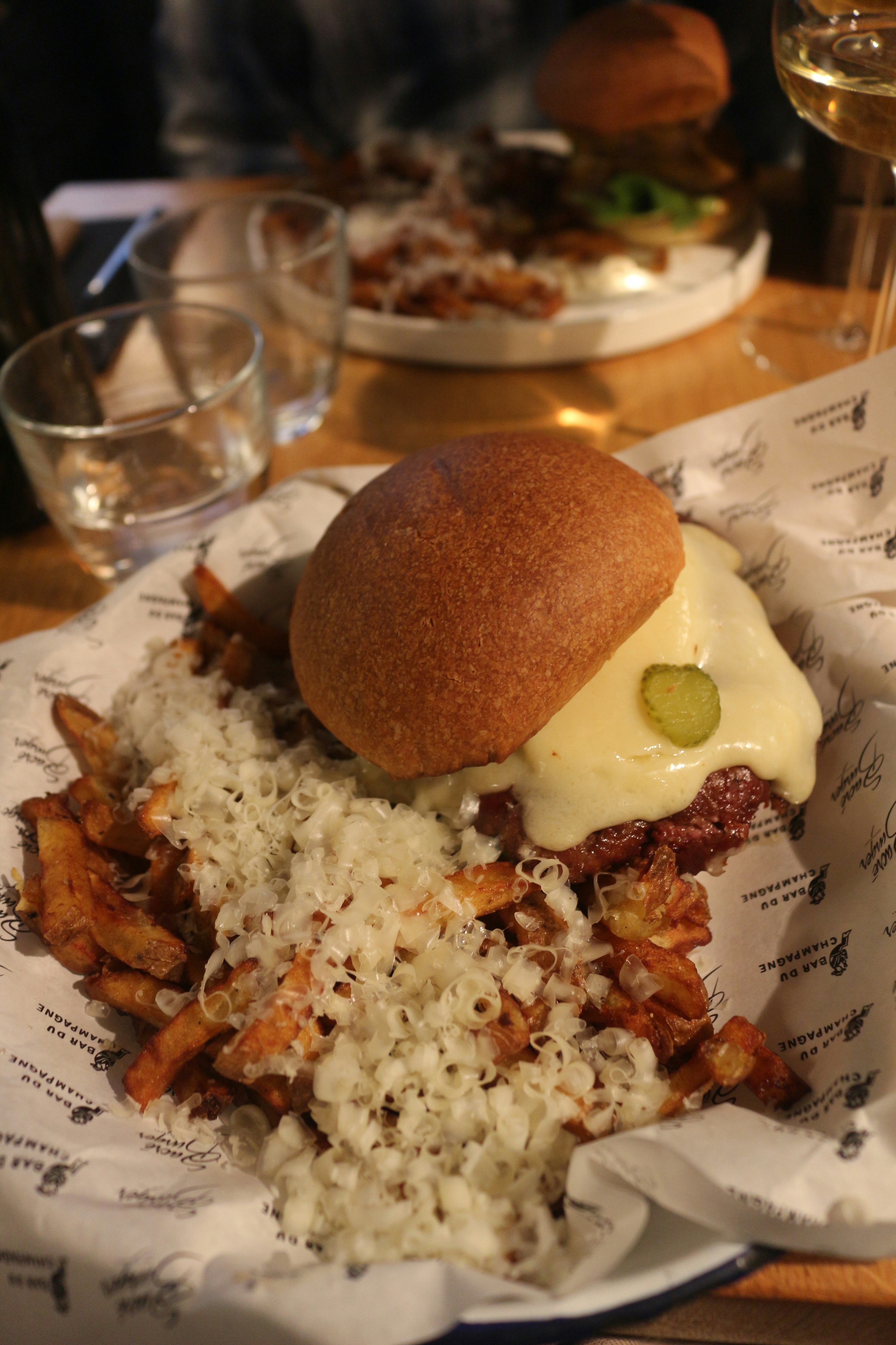 A juicy cheeseburger topped with pickles on a toasted bun, served alongside crispy fries covered with a generous amount of finely shredded cheese. The meal is presented on a piece of branded paper in a restaurant setting with a soft, warm light ambiance. A glass of wine and another plate with similar food are visible in the background.